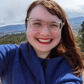 Emily Cordeaux, seen from the chest up, wearing a blue knit sweater, glasses, long brown hair, and smiling