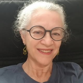 Alies Maybee, seen from the shoulders up, wearing a grey shirt, glasses, and smiling
