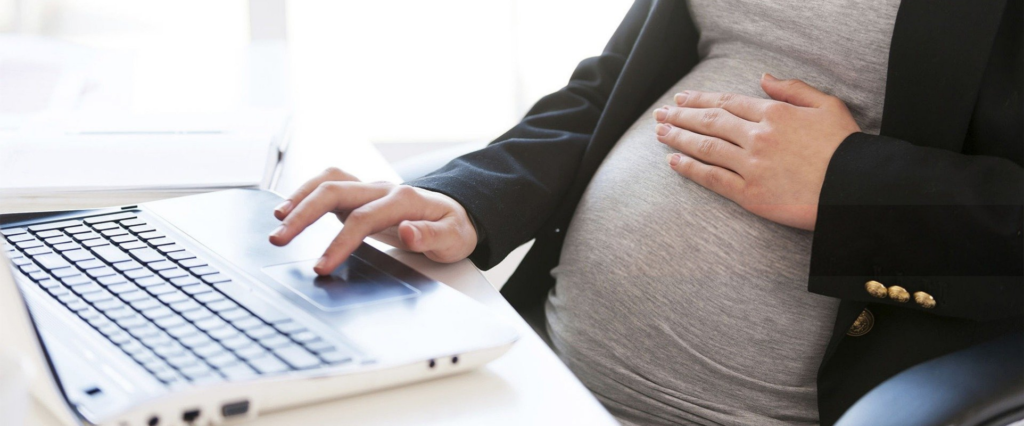 a pregnant women, sitting in a chair, is seen from the chest down with one hand on her belly and one on the trackpad of a laptop