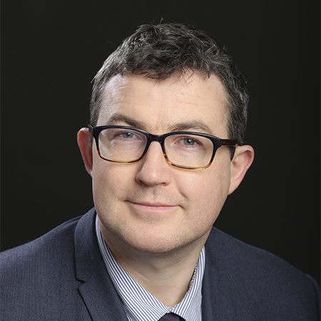 Owen Lyons, seen from the shoulders up, wearing a suit, glasses, short grey and black hair. Smiling